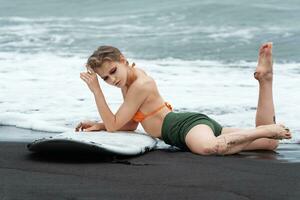 Dreamy sexy surfer is lying on black sandy beach with surfboard, showing off fit and athletic body photo