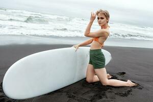 Female surfer kneeling on black sand of beach, holding her surfboard in hands and looking at camera photo