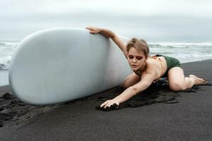 Female surfer kneels on beach, stretching forward with back arched, holding surfboard, looking down photo