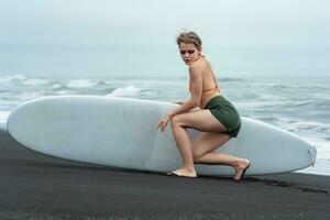 Female sports fashion model holding surfboard and sits in front of it, thoughtfully looking down photo