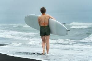 Rear view female surfer walking ankle deep water on beach carrying surfboard during summer vacation photo