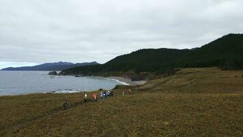Top view of tourists walking along mountain coast trail. Clip. Hikers walk along mountain coast road on cloudy day. Tourists walk along northern mountain coast on background of sea and forests photo