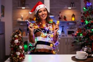 Happy young woman trying to decorate tree with lights on string for christmas holiday in festive kitchen at home. Caucasian smiling person knotted in bright tangled garland wire bulbs photo