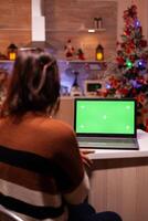 Caucasian young woman looking at green screen on laptop digital device sitting at festive home. Adult using modern chroma key notebook gadget on background display for mockup template photo