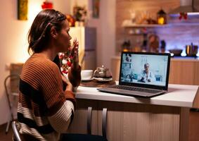 Woman with sickness calling doctor on online video conference while sitting in festive kitchen at home. Ill young patient using telemedicine for healing treatment and healthcare advice photo