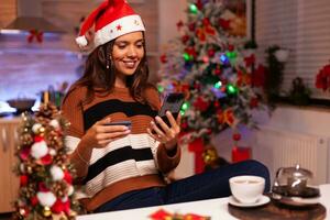 joven mujer pago con crédito tarjeta en compras sitio utilizando teléfono inteligente a hogar. caucásico adulto mecanografía número información, ordenando Navidad regalos en línea para amigos a celebracion foto