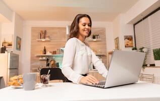 Businesswoman doing overtime working on laptop in home kit to grow business. Concentrated smiling entrepreneur in home kitchen using notebook during late hours in the evening. photo