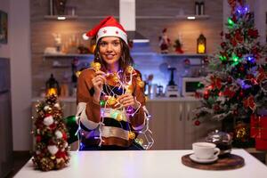 Caucasian woman decorating christmas tree with lights on string for holiday in festive kitchen at home. Young smiling person knotted in bright tangled garland wire illuminated bulbs photo