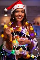 Cheerful adult tangled in christmas string lights used for decorating home. Festive caucasian young woman using garland with illuminated bulbs for seasonal decor and winter tree ornaments photo