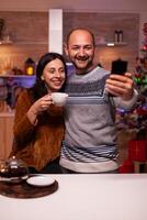 sonriente familia tomando selfie utilizando teléfono inteligente disfrutando Navidad hora en pie en Navidad cocina. contento alegre familia celebrando invierno fiesta estación. nuevo año festivo temporada foto