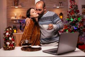 Happy boyfriend kissing girlfriend while discussing with remote friends during online videocall meeting conference using laptop. Cheerful family smiling enjoying christmas holiday together photo