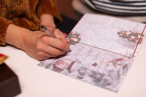 Closeup of woman hands writing xmas greeting card for family during christmas holiday standing in xmas decorated kitchen. Happy family enjoying spending winter season together photo