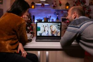 alegre familia hablando con remoto amigos durante en línea videollamada reunión en ordenador portátil computadora celebrando tiempo de Navidad juntos en Navidad decorado cocina. contento Pareja disfrutando invierno temporada foto