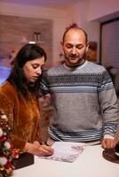 Cheerful girlfriend writing christmas greeting postcard celebrating xmas winter season with boyfriend standing in x-mas decorated kitchen, Happy family enjoying spending christmastime together photo