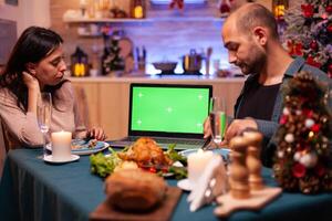 Happy family looking at green screen mock up chroma key tablet with isolated display standing on xmas decorated table. Couple celebrating christmas holiday enjoying winter season together photo