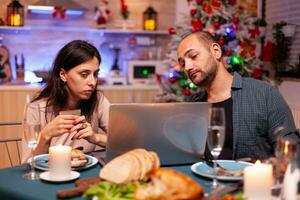 Cheerful couple shopping online xmas gift present paying with credit card on laptop computer sitting at christmas table in x-mas decorated kitchen. Happy family celebrating winter holiday photo