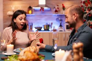 Haapy man proposing girlfriend to marry him using diamond luxury expensive engagement ring celebrating christmas holiday enjoying winter season together. Cheerful couple sitting at xmas dinning table photo