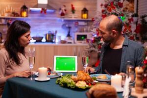 Cheerful family enjoying xmas dinner in x-mas decorated kitchen celebrating christmas holiday. Green screen mock up chroma key tablet computer with isolated display standing on table photo