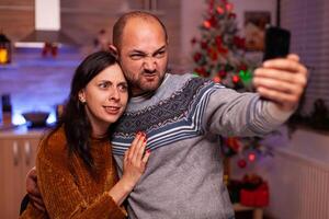 divertido Pareja tomando selfie utilizando teléfono inteligente mientras celebrando Navidad fiesta en pie en Navidad decorado cocina. contento alegre familia disfrutando gasto hora juntos durante invierno temporada foto