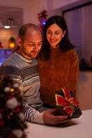 Husband surprising girlfriend with xmas present gift with ribbon on it celebrating christmas holiday in x-mas decorated kitchen. Happy joyful family enjoying winter season together photo