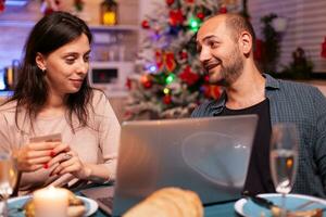 Cheerful couple buying xmas gift present paying online with credit card on laptop computer sitting at dining table in x-mas decorated kitchen. Happy family shopping for winter holidays photo