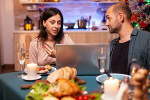 Cheerful couple buying xmas gift present paying online with credit card on laptop computer sitting at dining table in x-mas decorated kitchen. Happy family shopping for winter holidays photo