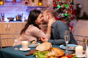 Happy couple kissing in xmas kitchen after marriage proposal spending christmas holiday sitting at dinning table. Joyful family enjoying engagement surprise celebrating winter season photo