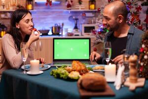 Happy family sitting at dining table in x-mas decorated kitchen enjoying winter season christmastime. Green screen mock up chroma key laptop computer with isolated display on table photo