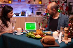 Happy family sitting at dining table in x-mas decorated kitchen enjoying winter season christmastime. Green screen mock up chroma key tablet computer with isolated display on table photo