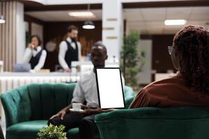 African american people at hotel using tablet with white screen, looking at blank template display and sitting in lounge area. Female guest holding device with isolated copyspace. photo