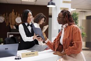 Young woman checking in at luxury hotel, filling in registration forms for records in lobby. African american guest doing check in with female receptionist at front desk, booking files. photo