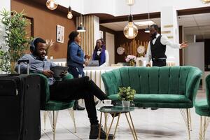 Smiling African American man traveler using tablet in hotel lounge area, black guy tourist wearing headphones sitting with suitcase in lobby listening to music while waiting for check-in process photo