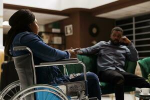 joven africano americano mujer turista con invalidez sentado en hotel vestíbulo con marido, llegando a silla de ruedas accesible hotel. sano hombre de viaje con discapacitado Novia foto