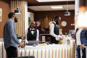 Pleasant conversation between receptionist and customer in hotel. Friendly smiling young African American woman front desk agent greeting guest at reception counter helping with check-in process photo