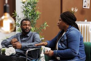 African American couple arriving at rehabilitation center for disabled, young man wheelchair user traveling with wife, sitting together in hotel lobby waiting for room with disability access photo