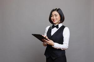 Smiling asian woman receptionist using digital tablet and looking at camera with positive facial expression. Catering service waitress in uniform standing with portable device portrait photo