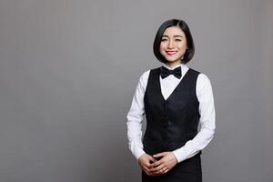 Attractive smiling asian receptionist wearing professional uniform standing and looking at camera with positive facial expression. Young waitress studio portrait on gray background photo