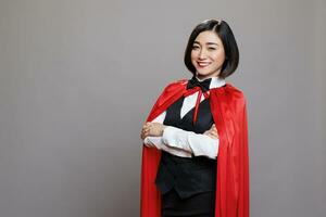 Confident asian waitress wearing hero red cloak, standing with folded hands and showing power portrait. Smiling woman receptionist in superwoman cape looking at camera in studio photo