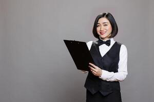 Attractive smiling asian waitress writing on a clipboard and looking at camera with positive facial expression. Cheerful young receptionist in uniform taking notes while posing in studio portrait photo