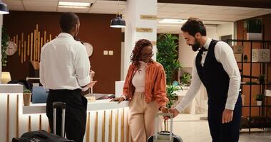 Happy relaxed hotel visitors ready to be checked in their room, receiving access key card from asian receptionist. Guests being assisted with their baggage by helpful professional porter photo