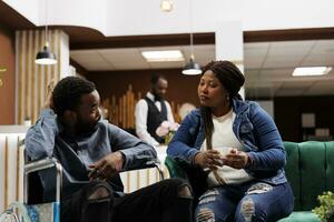 Handicapped travel. Young African American man on wheelchair waiting with wife in hotel lobby, guest with mobility impairments, traveling with disability. Travel companions for disabled travelers photo