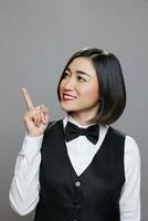 Smiling asian woman receptionist showing upwards and looking to side while advertising product. Cafe cheerful waitress wearing professional uniform pointing up with finger photo
