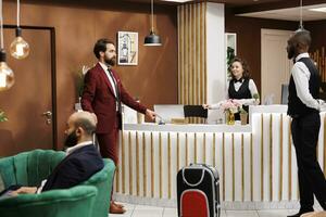 Front desk staff greets businessman with suitcase baggage, travelling on work trip and staying at luxury hotel. Formal person entering lobby to ask about room reservation, registration. photo