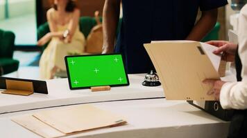 Guest asking about greenscreen info, pointing at tablet on reception counter in hotel lobby. Receptionist talking to traveller about isolated display mockup template, front desk concierge service. photo