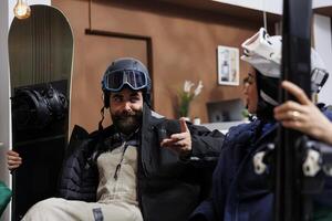 Youthful pair outfitted in cold-weather attire seated on sofa situated in hotel lobby engaged in conversation. Happy man and lady are clutching their wintersports gear set for snowy escapade. photo