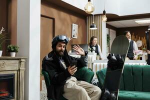 Caucasian male guest seated on sofa surrounded by snowboarding gear, having a video call on his digital phone. Tourist in snow clothing on videoconference with mobile device in ski resort lounge area. photo