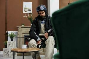 Caucasian male skier in winter clothing fine-tuning his snow equipment in ski resort lounge area, ready for snowy adventure. Youthful man seated near fireplace in hotel lobby is securing skiing skis. photo