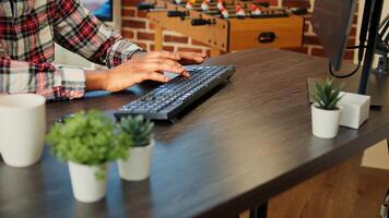 Employee remotely working from home on computer, inputing data in his brick wall apartment. African american freelancer typing on keyboard, teleworking at nice clean wooden desk, close up photo