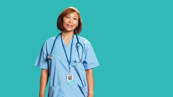 Happy proficient BIPOC nurse smiling while looking at camera. Portrait of cheerful asian healthcare specialist wearing professional scrubs, isolated over blue studio background photo