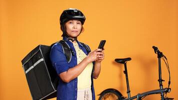 Portrait of courier waiting, looking in phone app for next customer order location. BIPOC woman delivering food with bycicle using thermic bag, isolated over studio background photo
